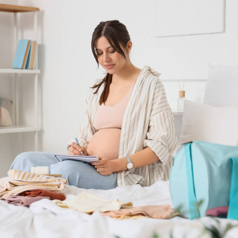 A pregnant woman sits on a bed surrounded by baby clothes, marking her checklist